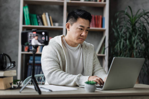 istock Pleasant young asian man working on laptop while recording video on his smart phone on tripod. 1446878676