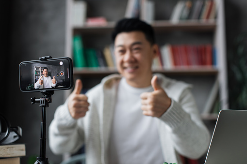 Focus on screen, attractive asian man in casual clothes talking and gesturing while recording video on modern phone on tripod showing thumbs up. Male blogger sitting at home and doing live stream.