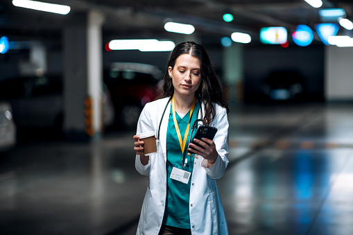 A young Caucasian female doctor is looking at her cellphone, while holding a cup of coffee and standing in an underground garage.