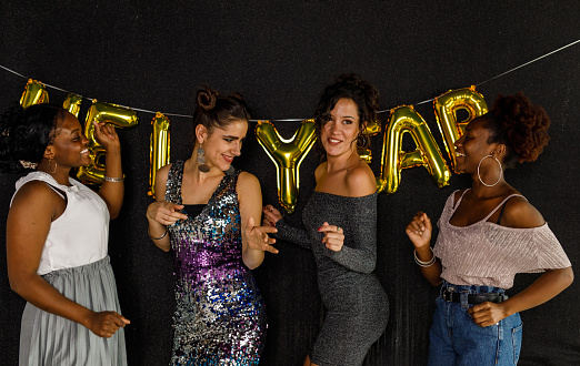 Candid shot of four diverse young women standing in front of a glittery black background, well dressed, dancing, bonding and having fun while enjoying the New Year's party celebration.