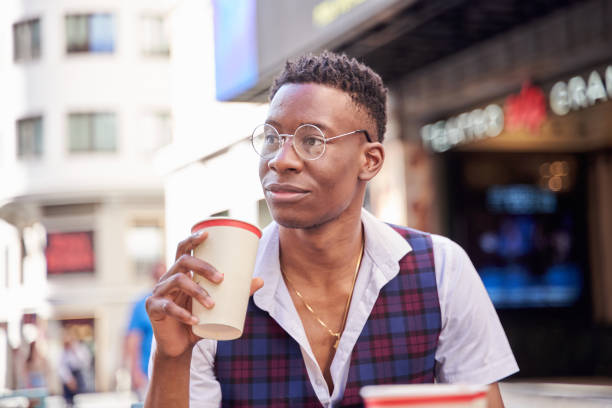 A blond adult woman in sunglasses smiling while holding a yellow coffee to-go cup outside in an outdoor seating area.