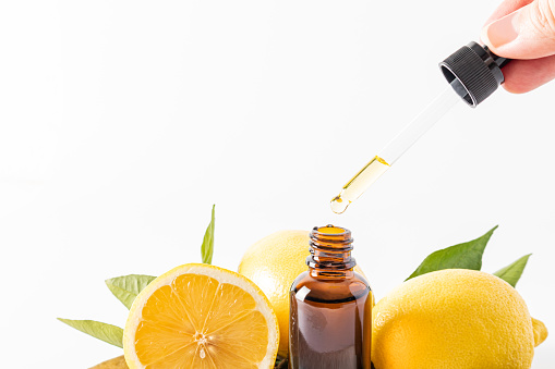 the fingers of a woman's hand hold a pipette with a lemon cosmetic over an open bottle of serum and lemons. Front view white background