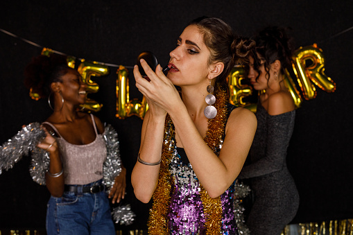 Candid shot of cool young woman standing on the dance floor, looking in the hand mirror and re-applying her lipstick during a New Year's eve party with friends. Her friends dancing and having fun in the background.