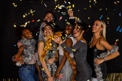 Candid shot of joyful group of diverse, young people, friends, standing on the dance floor, laughing, bonding, having fun playing with shiny tinsel garlands and dancing during a New Year's countdown while colorful confetti are falling down on them.