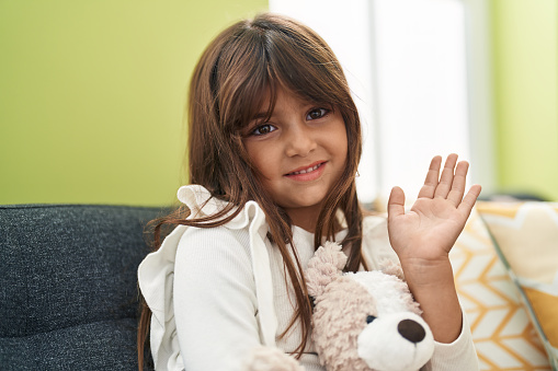 Adorable hispanic girl saying hello with hand hugging teddy bear at home
