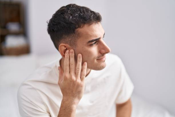 Young hispanic man suffering for ear pain sitting on bed at bedroom