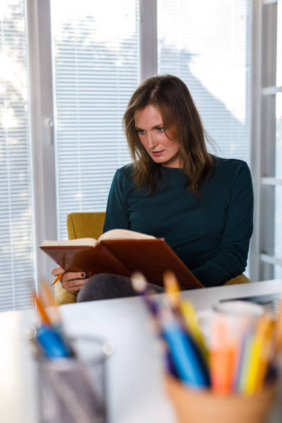 Portrait of diligent mid adult woman sitting at her desk at the office and reading her notes in a notebook while working on a project.