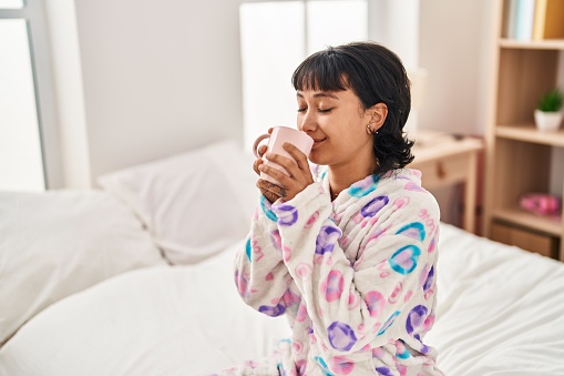 Young beautiful hispanic woman drinking cup of coffee sitting on bed at bedroom