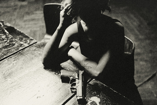 Portrait of a young woman with a pistol. She is sitting at a table. Her face is in shadow. A pistol lies in front of her in the faint light of an invisible ceiling light. It is the moment of deciding what to do next. Dark and mysterious.