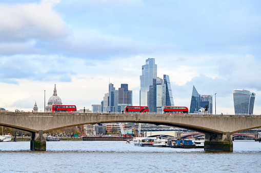 Side view over River Thames of double-decker buses and pedestrians on box girder bridge with historic City of London financial district in background.