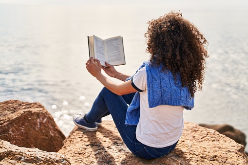 Young hispanic woman reading book sitting on rock at seaside