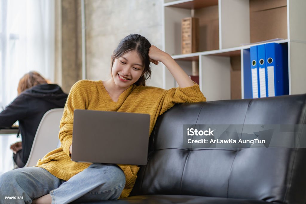 Young Asian woman listening to music on the sofa in the living room at home. Happy woman using laptop to video call with friends on vacation Adult Stock Photo