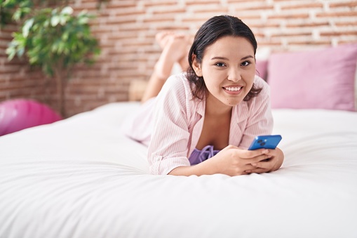 Young hispanic woman using smartphone lying on bed at bedroom