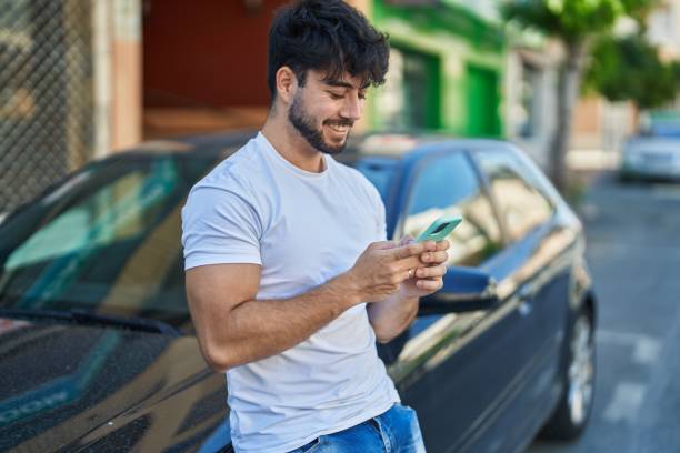 Young hispanic man using smartphone leaning on car at street