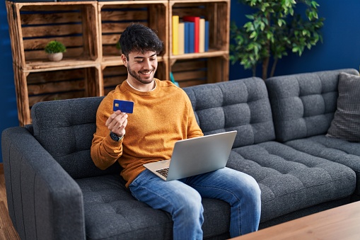 Young hispanic man using laptop and credit card sitting on sofa at home