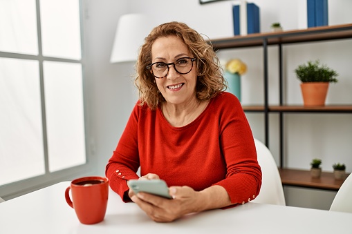 Middle age caucasian woman using smartphone and drinking coffee sitting on the sofa at home.