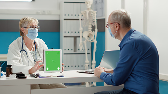 General practitioner and patient with face mask looking at greenscreen on digital tablet at checkup visit. Isolated chroma key template with blank copyspace and mockup background. Tripod shot.