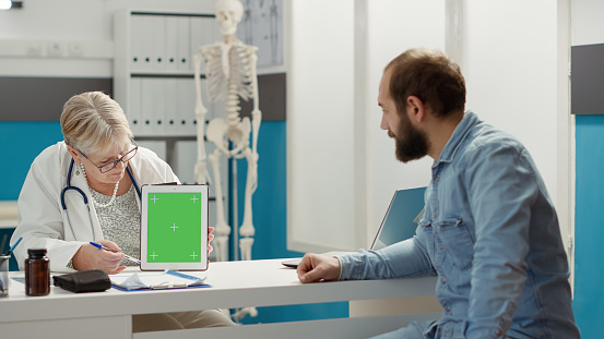Physician and patient analyzing digital tablet with greenscreen in medical office. Man and medic using blank copyspace mockup with isolated chromakey background and template. Tripod shot.