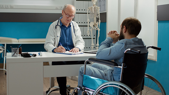 Health specialist doing consultation with wheelchair user in disability friendly cabinet. Medic examining patient with health condition and impairment, using expertise to talk to paralyzed man.