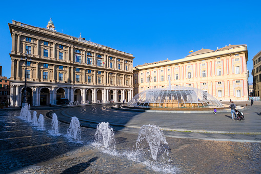 Tourists visiting Piazza De Ferrari in Genoa, the main city square characterized by a large circular fountain and many historic buildings, such as the Palazzo Ducale and the Palazzo della Regione