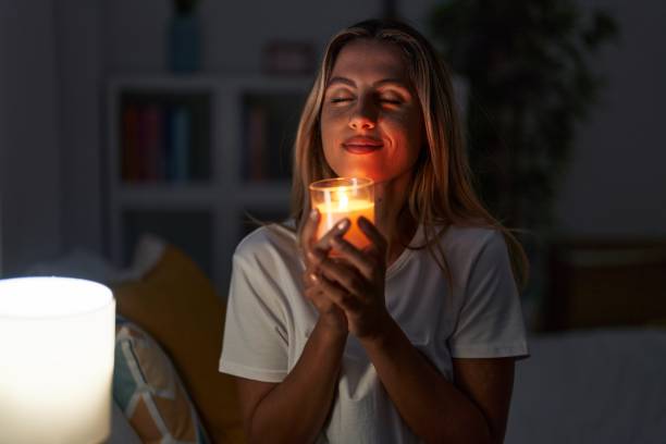 Young blonde woman sitting on bed smelling aromatic candle at bedroom Young blonde woman sitting on bed smelling aromatic candle at bedroom scented stock pictures, royalty-free photos & images