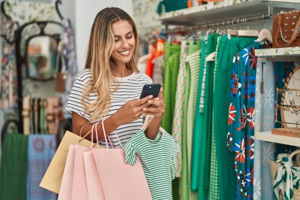 junge blonde kundin lächelt selbstbewusst mit smartphone im bekleidungsgeschäft - nur junge frauen stock-fotos und bilder