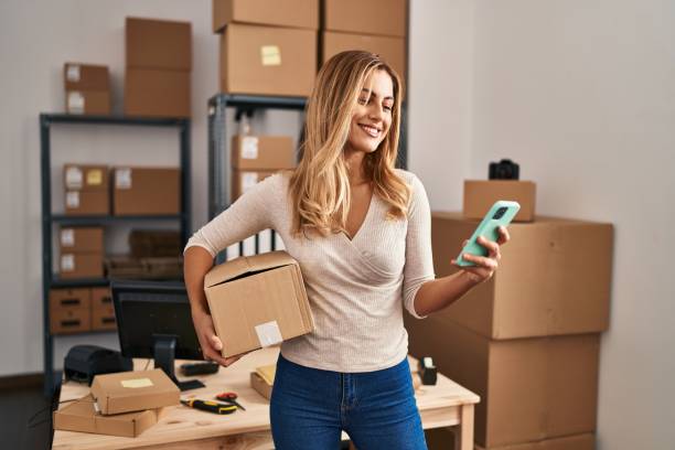 Young blonde woman ecommerce business worker using smartphone holding package at office