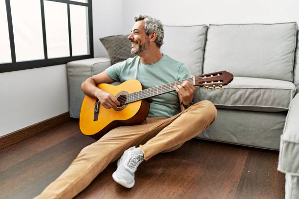 istock Middle age grey-haired man playing classical guitar sitting on floor at home 1446909609