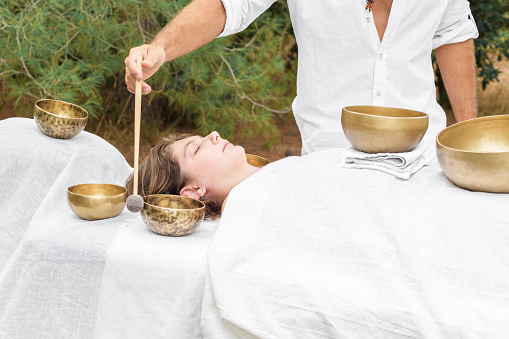 Young woman laying on a massage bed with tibetan singing bowl outdoor. A master of sound massage therapy doing a Buddhist healing practices, male hand holding a felt clapper