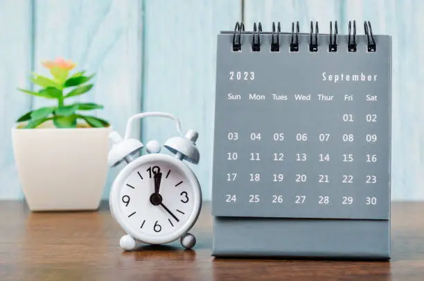 Photo of The September 2023 Monthly desk calendar for 2023 year with alarm clock on blue wooden background.
