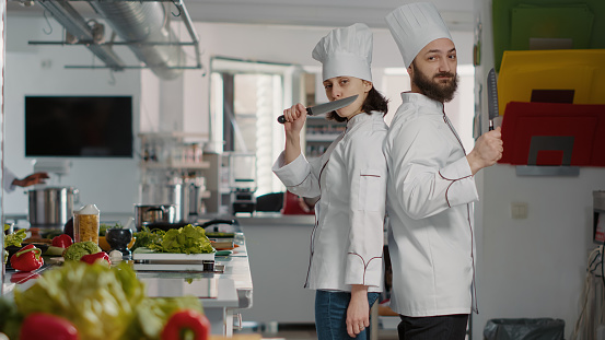 Portrait of chefs team acting funny with knives in restaurant kitchen, making joke about professional cuisine service. Cheerful man and woman working on gourmet meal dish with culinary recipe.