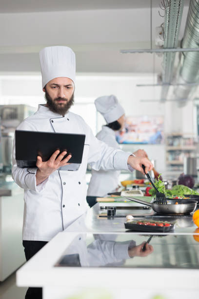 istock Chef preparing fresh organic vegetable garnish using pan and spatula while looking at laptop screen. 1446919521