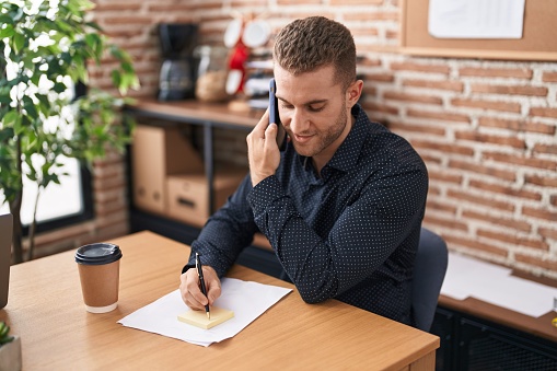 Young caucasian man business worker talking on smartphone writing on reminder paper at office
