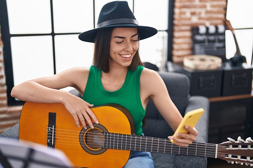 Young hispanic woman musician using smartphone playing classical guitar at music studio