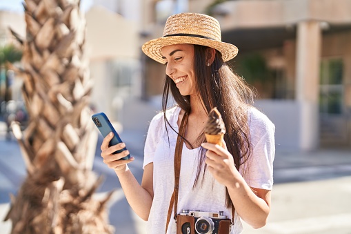 Young hispanic woman tourist using smartphone eating ice cream at street