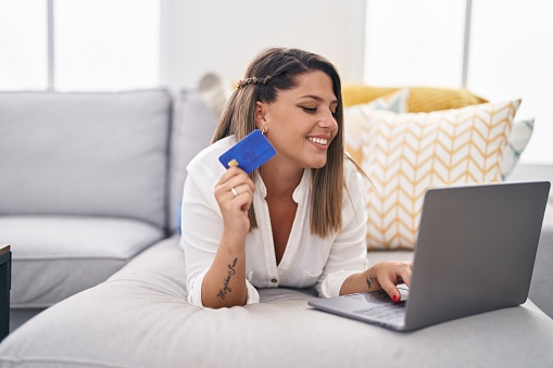 Young hispanic woman using laptop and credit card lying on sofa at home