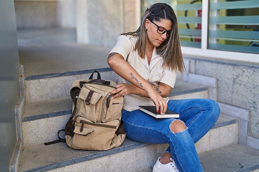 Young hispanic woman student putting notebook and book on backpack at university