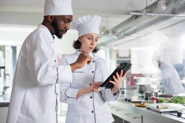 istock Professional chefs with wireless touchscreen device preparing ingredients for food contest held at restaurant. 1446923374