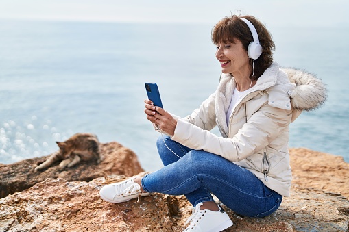Middle age woman listening to music sitting on the rock at seaside