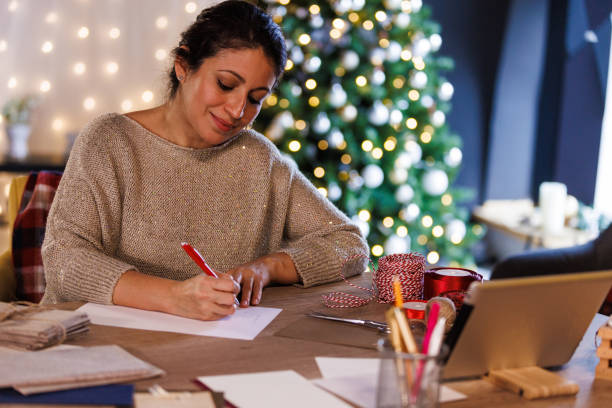 happy woman sitting at the table, writing christmas cards for loved ones - writing letter correspondence women imagens e fotografias de stock