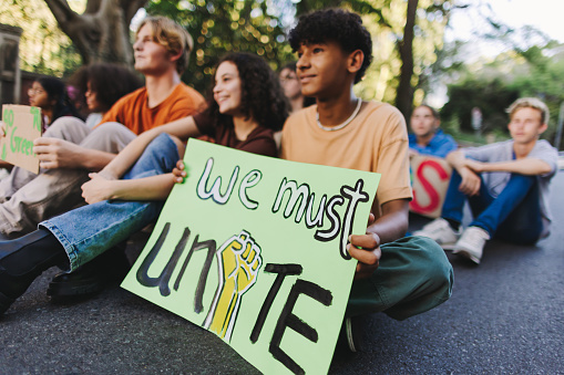 Teenage boy holding a poster while sitting with a group of youth demonstrators at a climate change protest. Multicultural young people joining the global climate strike.