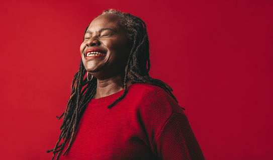 Elegant woman with dreadlocks smiling joyfully while standing against a red background. Happy mature woman embracing her natural hair with pride.