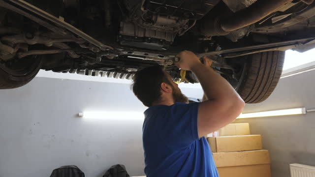 Camera moves to professional bearded auto mechanic repairing car with special tool at workshop. Young repairman in uniform working underneath a lifting vehicle at garage. Concept of automobile service.