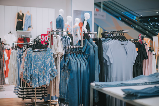 Closeup of smiling blond woman choosing clothes at department store  in local supermarket. She's holding a beige blouse and looking at it. The woman is wearing gray cap, red sweater and scarf. Side view.