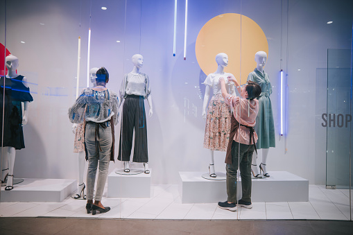 Asian female retail store worker display artist dressing up mannequin in store window display at womenswear clothing store