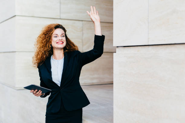 istock Smiling happy businesswoman dressed formally, holding in hands modern tablet computer, waving with her hand while noticing her companion, being glad to see him. Woman with curly hair saying `hello` 1446947656