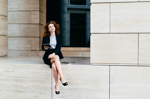 Lovely curly woman with confident look, sitting alone, demonstrating her slender long legs, dressed in black jacket with skirt, holding tablet, looking schedules or diagrams, making business report
