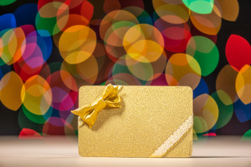 A close-up of a gold gift card with gold ribbon and a bow against a multi-colored bokeh background.