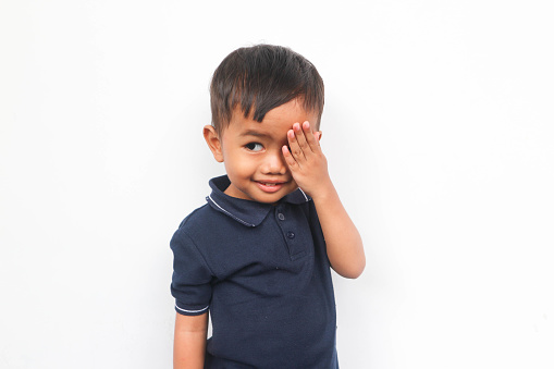 Cute little Asian boy shy, closing one eye with his fingers isolated on white background
