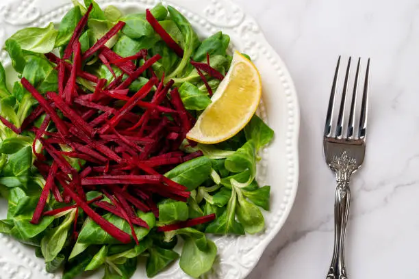 Lambs lettuce with raw beet vitamin salad. Grated raw beet over fresh cornsalad leaves and lemon slice on a white plate over marble surface. Vegan vegetable salad for low calories slimming diet. Top view.